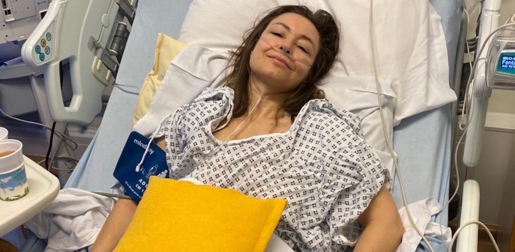 Living kidney donor Julie lying in a hospital bed. She is dressed in a hospital gown with her head laying against the pillow, and is smiling at the camera