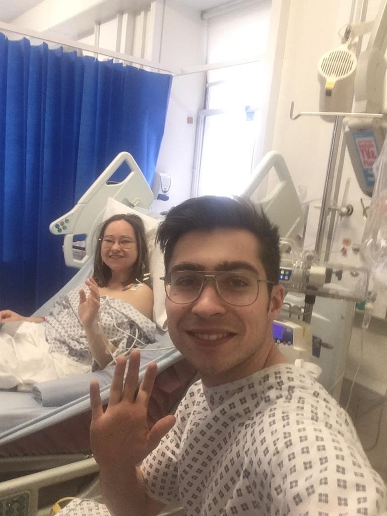 Living kidney donor Henry and his sister and recipient. The two are in hospital and are both wearing hospital gowns. Henry is taking a selfie of them both smiling and waving for the camera.