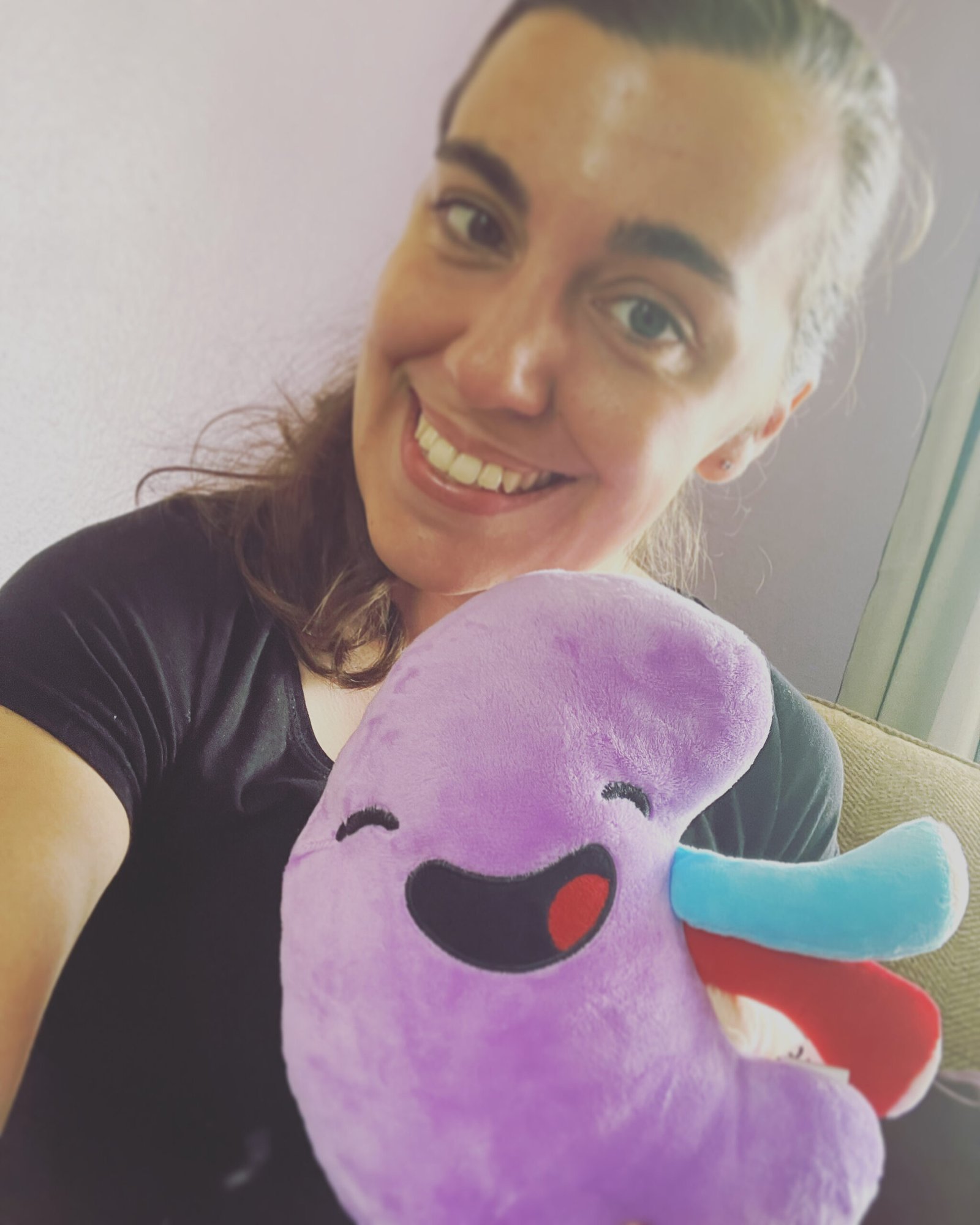 Living kidney donor Georgia. She is taking a selfie of herself and smiling at the camera, while holding a smiling stuffed kidney plushie