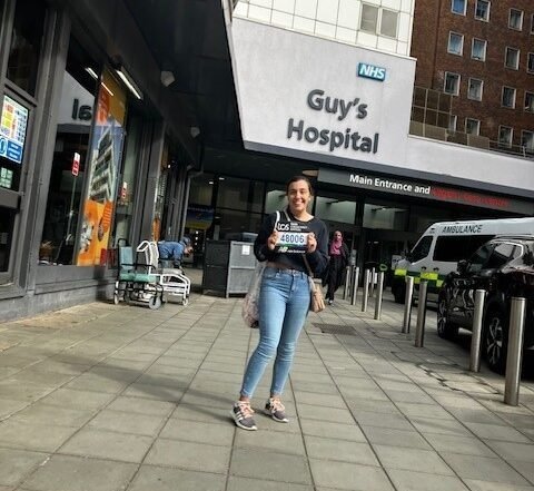 Living kidney donor Georgia standing outside of Guy's Hospital, where her transplant took place. She is standing some distance from the photographer and smiling towards the camera, with the entrance to Guy's Hospital behind her.