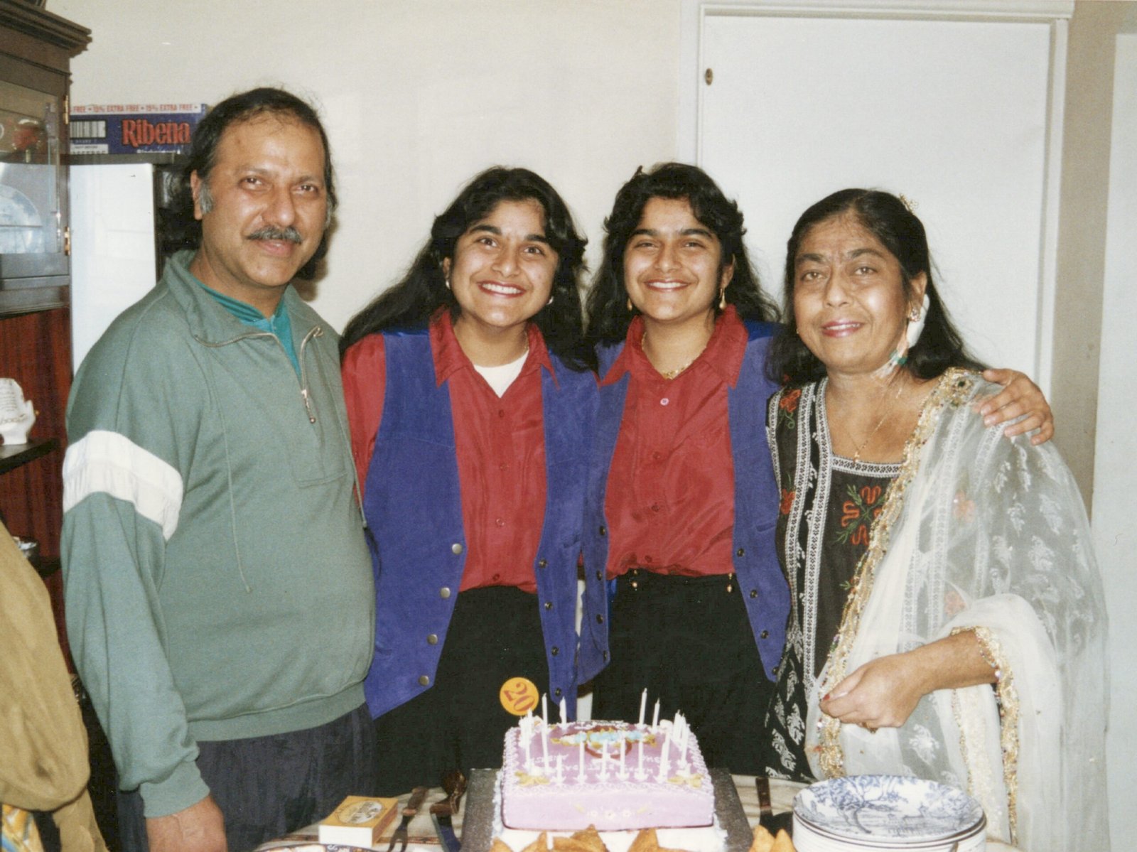 A young living kidney donor Aisha alongside her sister, mum and dad. The two sisters are in matching blue waistcoats with a red shirt underneath. All four are standing in front of a white wall and are smiling towards the camera.