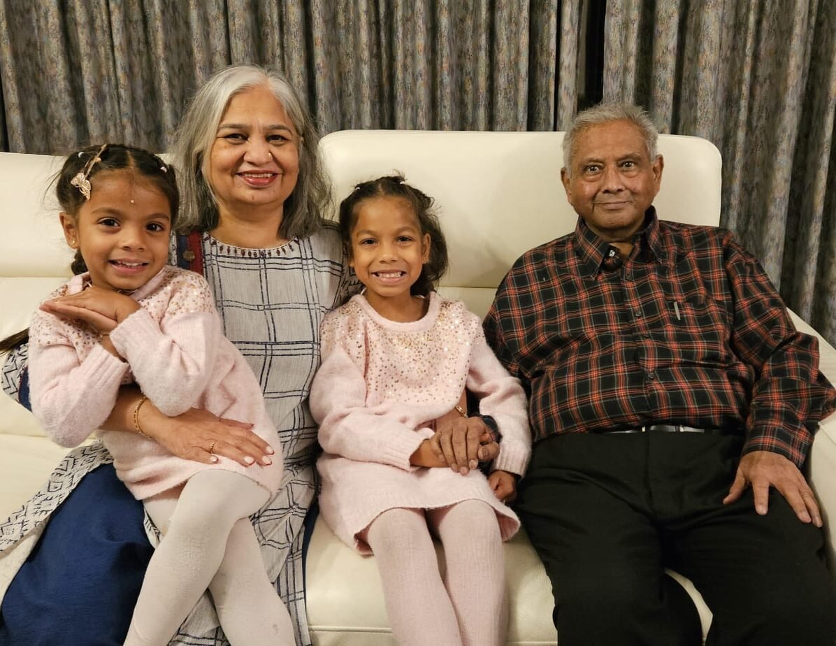 Image of living kidney donor Harsha alongside her husband and living kidney recipient Kirit. Harsha has two of their young granddaughters sitting on her lap, both wearing pink dresses and smiling for the camera.