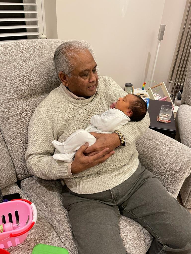Image of Harsha's husband and kidney recipient Kirit, sitting on a sofa andholding his newborn granddaughter