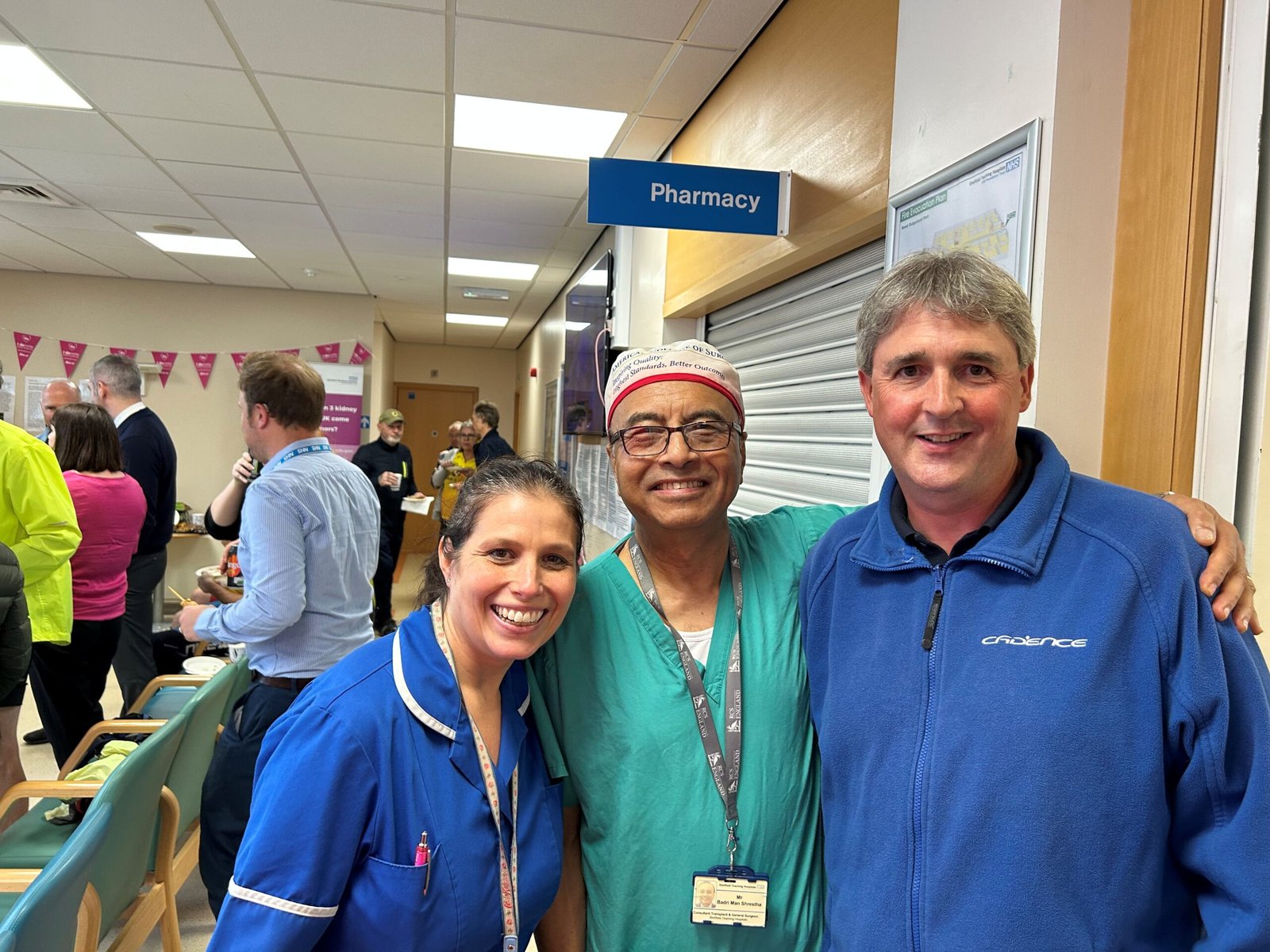 Image of living kidney donor Leigh in a hospital hallway alongside his surgeon and another healthcare professional, all smiling widely for the camera.