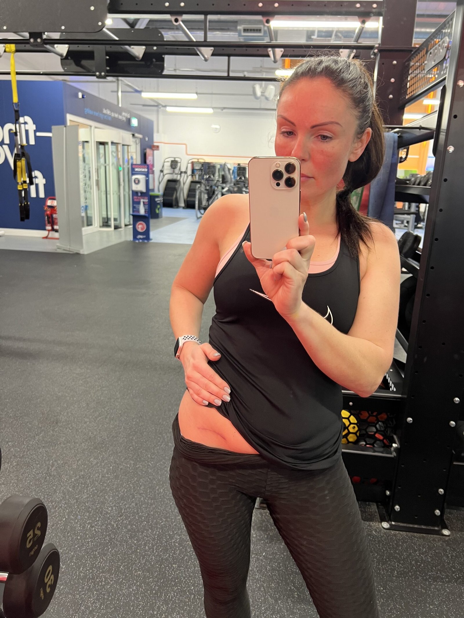 Image of living kidney donor Marilyse taking a selfie in a mirror at a gym. She is wearing black gym clothes and is lifting her top to show a small scar on her waist where her kidney was taken out.