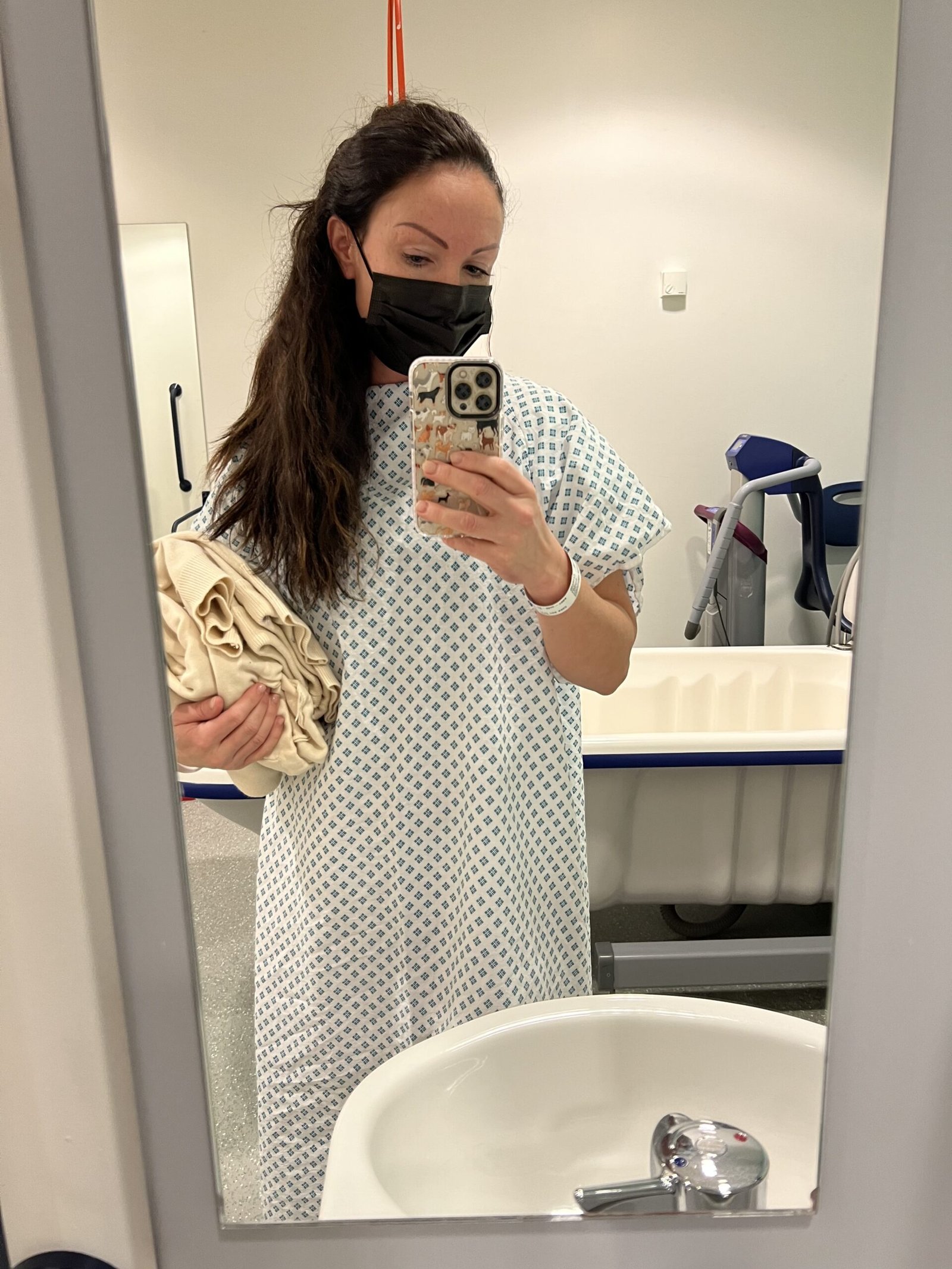 Image of living kidney donor Marilyse in hospital, wearing a hospital gown and facemask, taking a selfie of herself in a mirror.