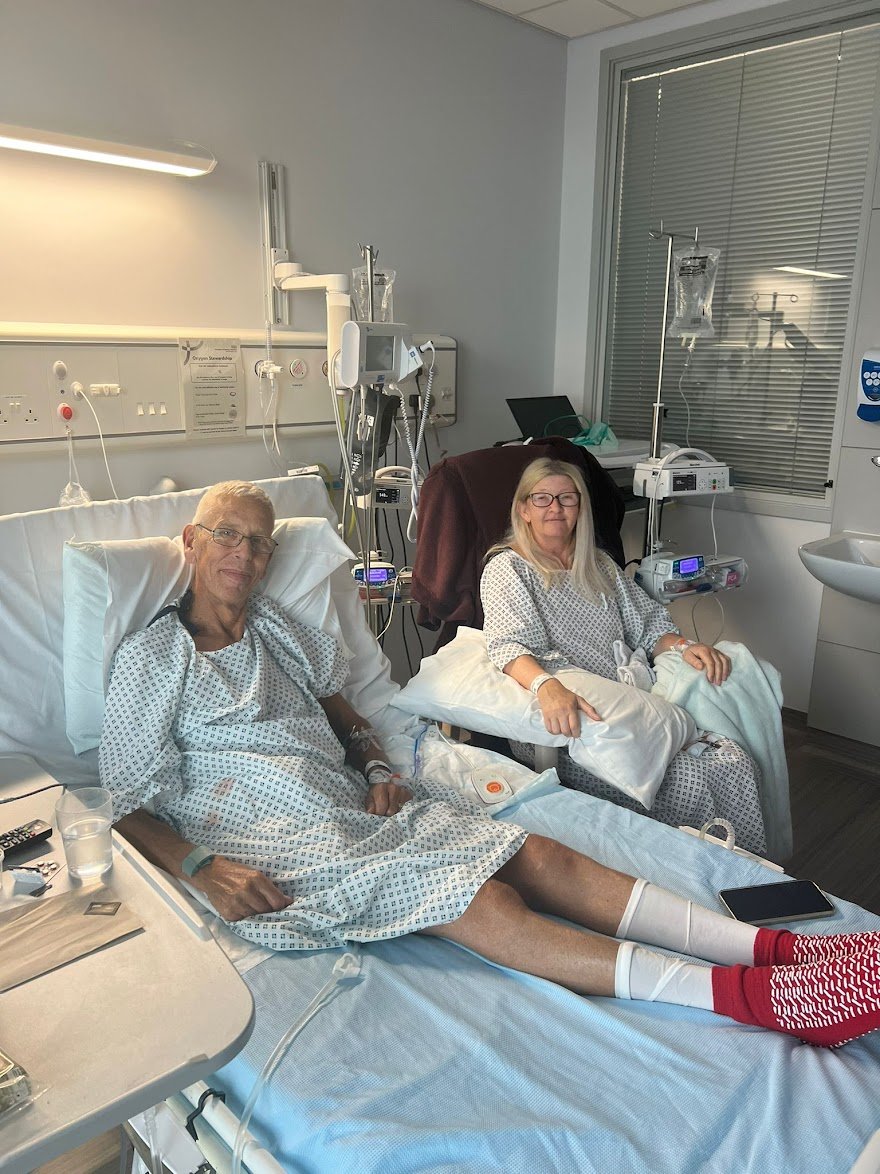Image of living kidney recipient Roy lying in a hospital bed. To his side is his wife Carolyn, sitting on a chair. They are both smiling warmly for the camera.
